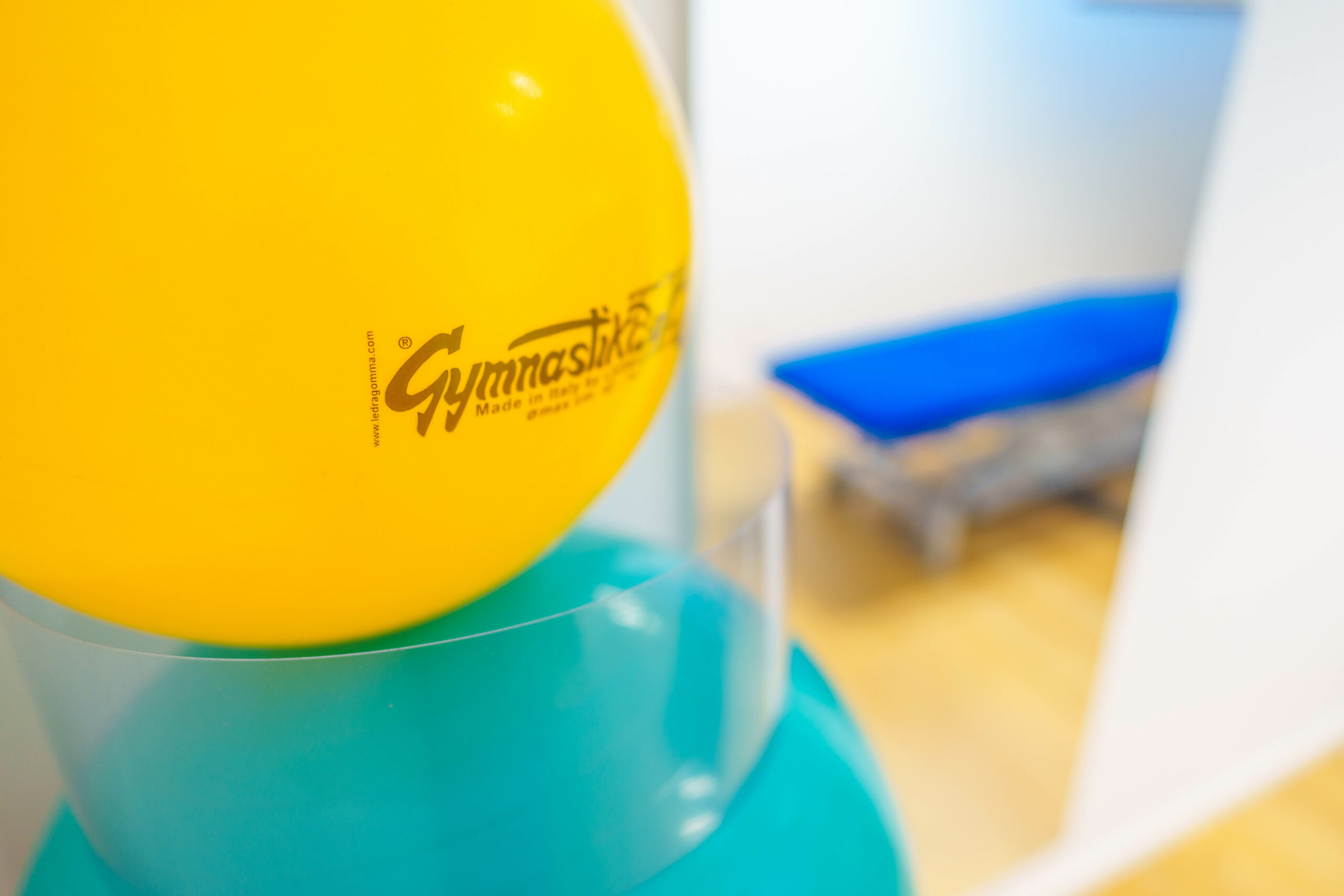 Gelber Gymnastikball im Vordergrund, mit unscharfem blauem Behandlungstisch im Hintergrund einer Physiotherapiepraxis.