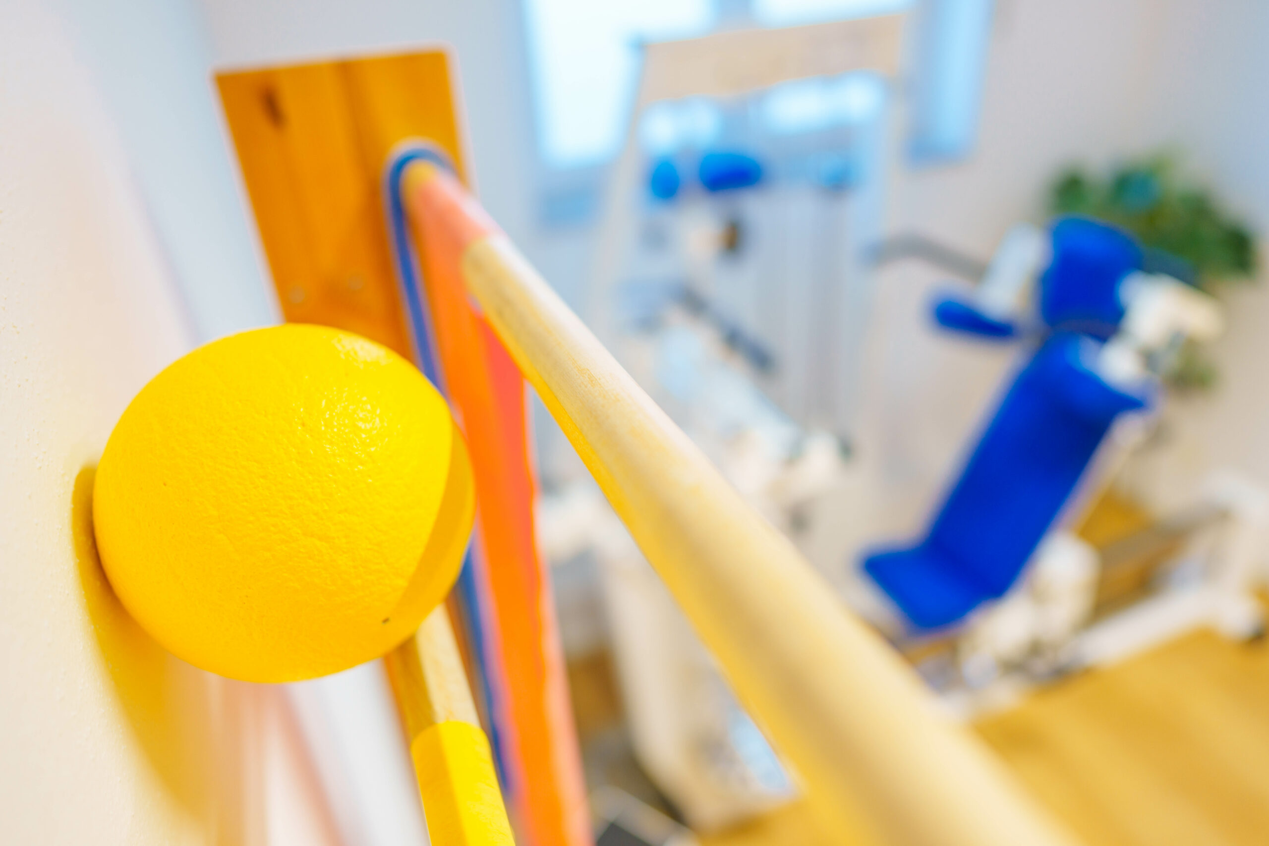 Nahaufnahme einer gelben Therapiekugel an einer Sprossenwand, mit Fitnessgerät und bunten Trainingsbändern im Hintergrund, in einem Physiotherapieraum.