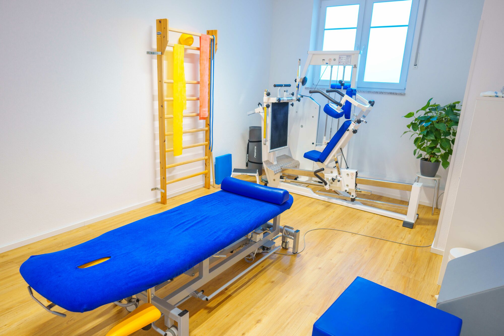 Physiotherapieraum mit blau bezogenem Behandlungstisch, Sprossenwand, Fitnessgerät und bunten Therapiebändern, ideal für Rehabilitations- und Trainingsprogramme.